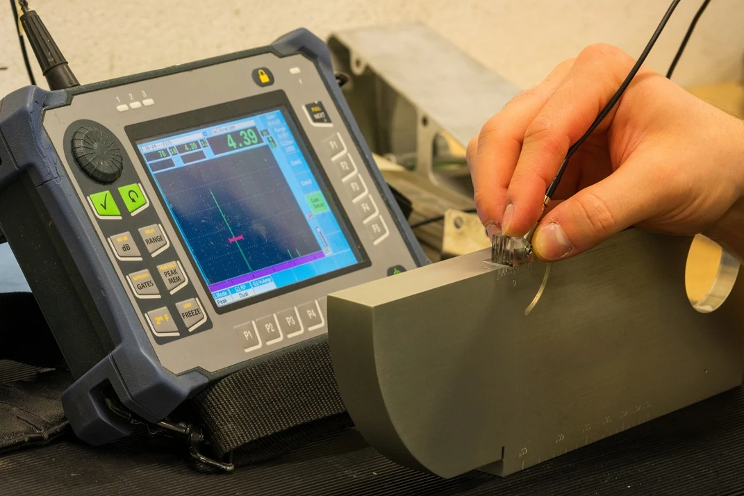  Technician performing ultrasonic NDT with a handheld flaw detector and contact probe on a machined calibration block; A‑scan visible on the screen. Inspected by APEC Inspection