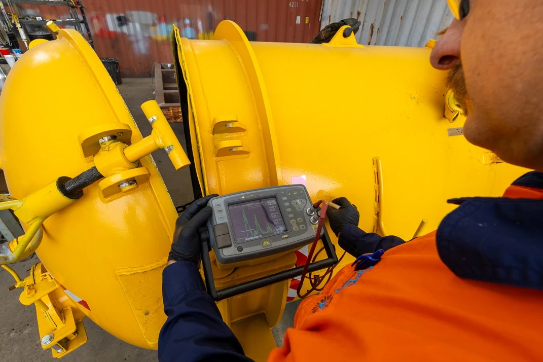 Technician performing ultrasonic inspection with a handheld flaw detector on a large yellow flanged metal component.