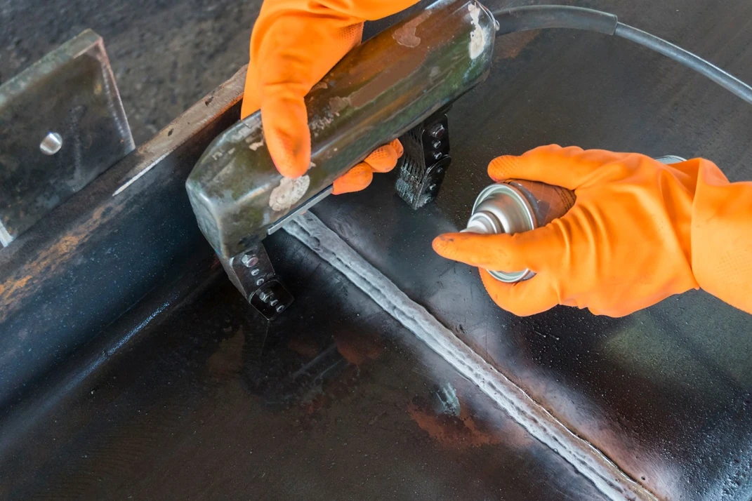  A close-up of hands wearing orange gloves, one holding a nozzle spray can and the other gripping a green tool, performing magnetic particle inspection on a metallic surface with a visible weld seam. Inspected by APEC Inspection
